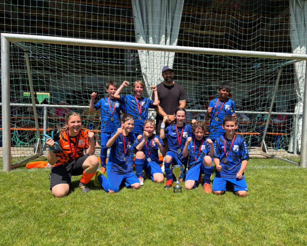 Die E-Jugend des TSV Wöschbach posiert nach dem Gewinn des Pfinztal-Cups vor einem Fußballtor. Die Kinder tragen blaue Trikots und Medaillen, jubeln gemeinsam mit ihrem Trainer und präsentieren stolz den Siegerpokal. Das Mannschaftsfoto entstand bei sonnigem Wetter auf dem Sportgelände in Söllingen.