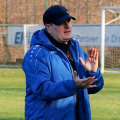 Trainer_Alexander_Heidt Alexander Heidt steht am Rand eines Fußballplatzes und trägt eine blaue Sportjacke sowie eine dunkle Baseballkappe. Die Person gestikuliert mit beiden Händen, als würde sie Anweisungen geben oder etwas erklären. Im Hintergrund sind ein Fußballtor, ein Zaun und kahle Bäume zu sehen. Die Aufnahme entstand im Freien bei Tageslicht.